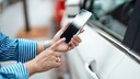 Eine Person h&auml;lt ein iPhone vor die Fahrert&uuml;r eines wei&szlig;en Autos und tippt auf den Bildschirm. Das Smartphone befindet sich direkt neben dem T&uuml;rgriff, im Hintergrund sind weitere Fahrzeuge unscharf zu sehen.