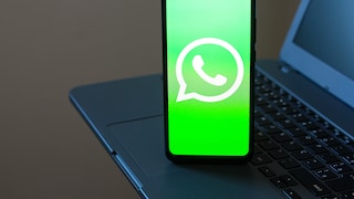 Ein Smartphone steht auf einer Laptop-Tastatur. Auf dem hellgrünen Display ist das weiße WhatsApp-Logo zu sehen. Im Hintergrund sind Teile des Laptops unscharf erkennbar.