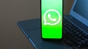 Ein Smartphone steht auf einer Laptop-Tastatur. Auf dem hellgr&uuml;nen Display ist das wei&szlig;e WhatsApp-Logo zu sehen. Im Hintergrund sind Teile des Laptops unscharf erkennbar.