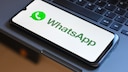 Smartphone mit ge&ouml;ffnetem WhatsApp-Startbildschirm liegt schr&auml;g auf der Tastatur eines Laptops. Auf dem Display sind das gr&uuml;ne WhatsApp-Logo und der Schriftzug &bdquo;WhatsApp&ldquo; auf wei&szlig;em Hintergrund zu sehen. Die schwarzen Tasten der Tastatur f&uuml;llen den Hintergrund des Bildes.