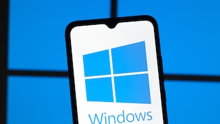 Smartphone mit groß dargestelltem Windows-Logo auf dem Bildschirm vor einem unscharfen blauen Hintergrund, der an die typischen Fenster des Windows-Logos erinnert. Das Gerät steht leicht schräg im Vordergrund, während die blaue Beleuchtung im Hintergrund das Windows-Motiv aufgreift.