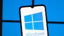 Smartphone mit gro&szlig; dargestelltem Windows-Logo auf dem Bildschirm vor einem unscharfen blauen Hintergrund, der an die typischen Fenster des Windows-Logos erinnert. Das Ger&auml;t steht leicht schr&auml;g im Vordergrund, w&auml;hrend die blaue Beleuchtung im Hintergrund das Windows-Motiv aufgreift.
