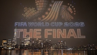 Alt-Text: Drohnenshow über einer nächtlichen Skyline am Wasser. Hunderte beleuchtete Drohnen formen in der Luft die Schriftzüge „FIFA WORLD CUP 2026“ und „THE FINAL“ sowie ein Fußballmotiv. Im Hintergrund sind zahlreiche beleuchtete Hochhäuser und deren Spiegelungen auf der Wasseroberfläche zu sehen.