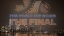 Alt-Text: Drohnenshow &uuml;ber einer n&auml;chtlichen Skyline am Wasser. Hunderte beleuchtete Drohnen formen in der Luft die Schriftz&uuml;ge &bdquo;FIFA WORLD CUP 2026&ldquo; und &bdquo;THE FINAL&ldquo; sowie ein Fu&szlig;ballmotiv. Im Hintergrund sind zahlreiche beleuchtete Hochh&auml;user und deren Spiegelungen auf der Wasseroberfl&auml;che zu sehen.