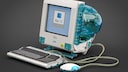 Lego-Nachbau eines Apple iMac G3 auf dunklem Hintergrund. Der Computer besitzt ein transparentes blaues Geh&auml;use mit integriertem Bildschirm, auf dem das Mac-OS-Logo zu sehen ist. Vor dem Rechner liegen eine passende Tastatur und die runde &bdquo;Hockey-Puck&ldquo;-Maus, die &uuml;ber transparente Kabel mit dem Modell verbunden sind.
