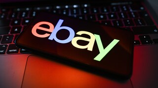 Ein Smartphone liegt schräg auf der Tastatur eines Laptops. Auf dem Display ist das farbige Ebay-Logo auf dunklem Hintergrund zu sehen. Die Tastatur und das Gehäuse des Laptops werden von roten und violetten Lichtreflexen beleuchtet. Der Hintergrund ist unscharf.