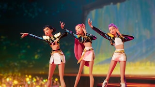 Drei animierte Figuren stehen auf einer beleuchteten Bühne und führen eine Choreografie auf. Alle drei tragen kurze weiße Outfits mit schwarzen, glitzernden Jacken und goldfarbenen Details. Die Figur links streckt einen Arm nach vorne aus, die mittlere Figur mit langen pinken Haaren hebt beide Arme an, die Figur rechts mit langen violetten Haaren hält eine Hand am Kopf. Im unscharfen Vordergrund sind Lichter und Publikum zu sehen. Der Hintergrund zeigt eine farbig beleuchtete Konzertkulisse in Blau-, Grün- und Gelbtönen.