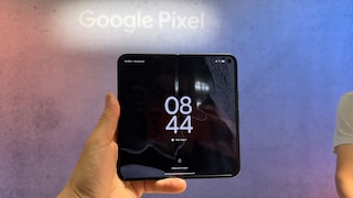 Eine Hand hält ein aufgeklapptes Google Pixel 11 Pro Fold vor einer großflächigen Hintergrundwand mit dem Schriftzug „Google Pixel“. Auf dem inneren Display des faltbaren Smartphones ist der Sperrbildschirm mit der Uhrzeit 08:44 zu sehen. Die Knickstelle in der Mitte des Displays ist sichtbar. Der Hintergrund ist in Rosa- und Blautönen gehalten, rechts ist ein angeschnittener Teil einer Person in hellem Oberteil zu erkennen.
