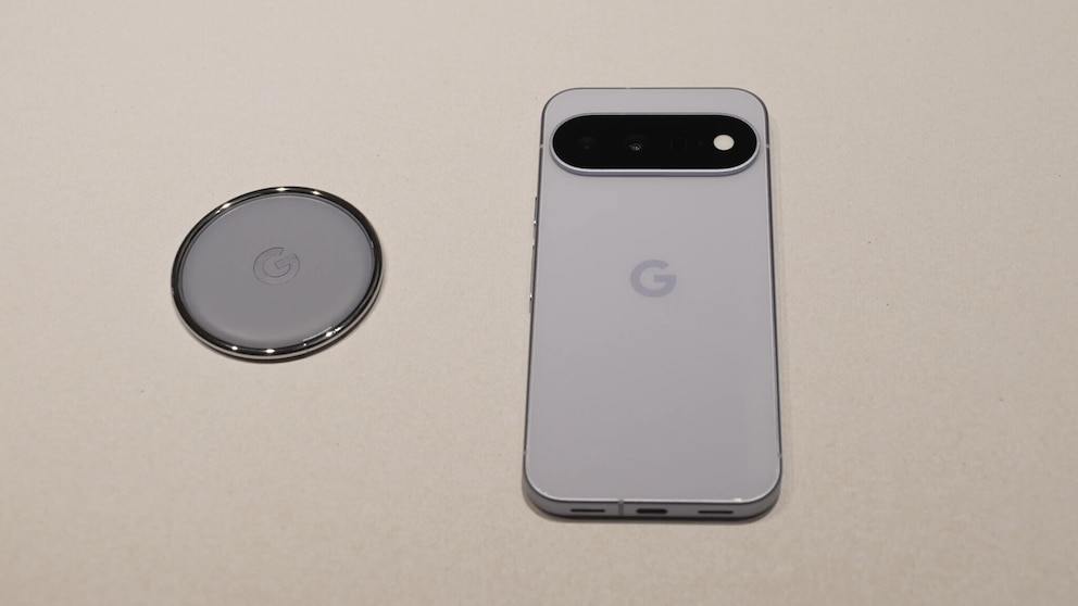 Ein Google Pixel 11 liegt auf einer hellen Oberfl&auml;che neben einem runden Pixel Tag. Zu sehen sind die R&uuml;ckseiten beider Ger&auml;te in heller Farbe. Das Smartphone verf&uuml;gt &uuml;ber ein schwarzes, horizontal verlaufendes Kameramodul im oberen Bereich. Der Pixel Tag ist kreisf&ouml;rmig und tr&auml;gt mittig das Google-Logo. Beide Ger&auml;te liegen nebeneinander und sind von oben fotografiert.