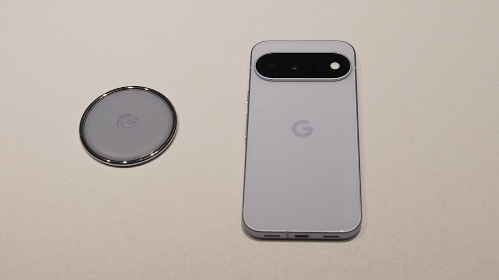 A Google Pixel 11 lies on a light surface next to a round Pixel Tag. The backs of both devices are visible in light color. The smartphone features a black, horizontally aligned camera module at the top. The Pixel Tag is circular and bears the Google logo in the center. Both devices are photographed from above.