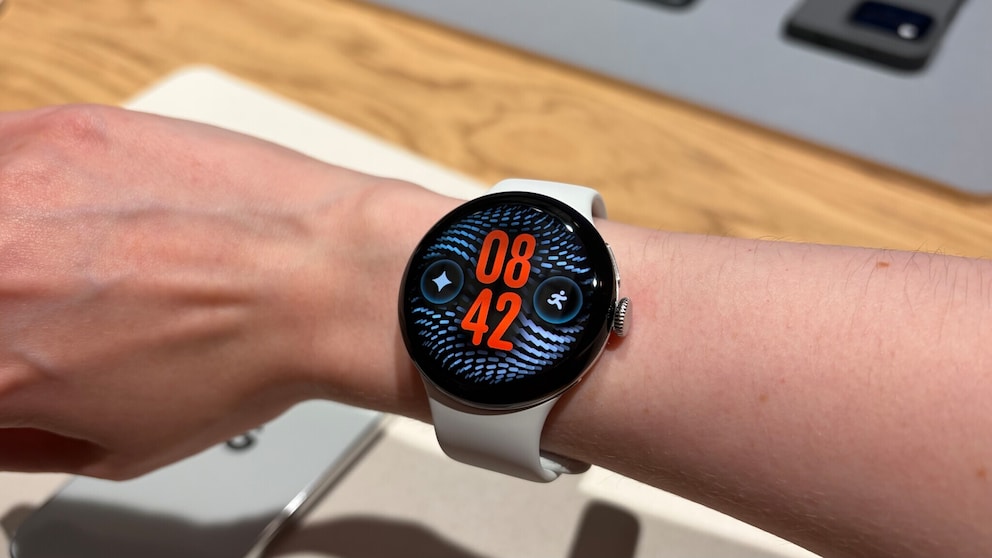 A person wears a Google Pixel Watch 5 with a light band on the wrist. The round display shows the time 08:42 in large red digits. Next to it are two symbols. The smartwatch is on a wooden table, with more devices in the blurred background.