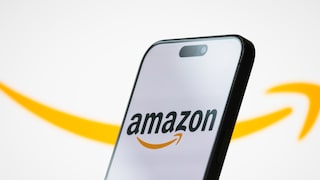 Schräg aufgestelltes Smartphone vor hellem Hintergrund. Auf dem Display ist das Amazon-Logo mit schwarzem Schriftzug und orangem Pfeil zu sehen. Im Hintergrund verläuft groß und unscharf der orangefarbene Amazon-Pfeil über das Bild. Das Smartphone besitzt einen schwarzen Rahmen und eine pillenförmige Aussparung am oberen Displayrand.