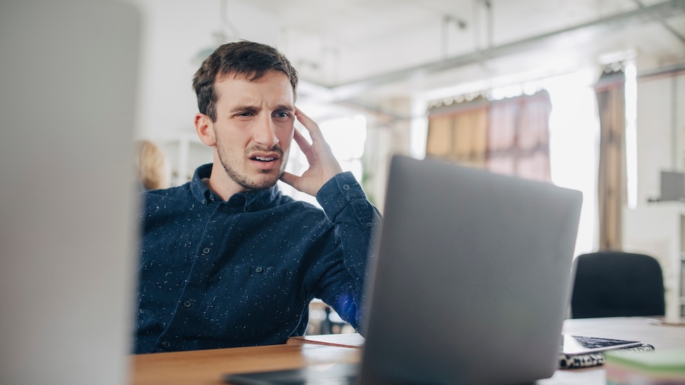 Eine Person sitzt an einem Schreibtisch vor einem aufgeklappten Laptop. Eine Hand liegt am Kopf. Auf dem Tisch stehen weitere Arbeitsgeräte, darunter ein zweiter Bildschirm im Vordergrund. Der Hintergrund zeigt einen hellen Büroraum mit Fenstern und unscharfen Möbeln.