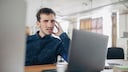 Eine Person sitzt an einem Schreibtisch vor einem aufgeklappten Laptop. Eine Hand liegt am Kopf. Auf dem Tisch stehen weitere Arbeitsger&auml;te, darunter ein zweiter Bildschirm im Vordergrund. Der Hintergrund zeigt einen hellen B&uuml;roraum mit Fenstern und unscharfen M&ouml;beln.