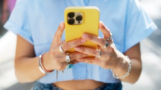 Eine Person hält ein gelbes Smartphone mit drei Kameralinsen auf der Rückseite in beiden Händen. Die Person trägt ein hellblaues Oberteil, mehrere Ringe an den Fingern sowie Armbänder an den Handgelenken. Der Hintergrund ist unscharf und hell ausgeleuchtet.