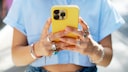 Eine Person h&auml;lt ein gelbes Smartphone mit drei Kameralinsen auf der R&uuml;ckseite in beiden H&auml;nden. Die Person tr&auml;gt ein hellblaues Oberteil, mehrere Ringe an den Fingern sowie Armb&auml;nder an den Handgelenken. Der Hintergrund ist unscharf und hell ausgeleuchtet.
