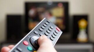 Eine Hand hält eine Fernbedienung in Richtung eines Fernsehers. Der Fernseher im Hintergrund ist unscharf, auf dem Bildschirm läuft eine nicht erkennbare Sendung. Links und rechts neben dem Fernseher stehen Lautsprecher.