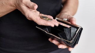 Eine Person hält eine geöffnete schwarze Geldbörse in der einen Hand und mehrere Euro-Münzen in der anderen. Im Geldbeutel befinden sich einige Geldscheine, das Münzfach wirkt weitgehend leer. Der Hintergrund ist unscharf und hell.