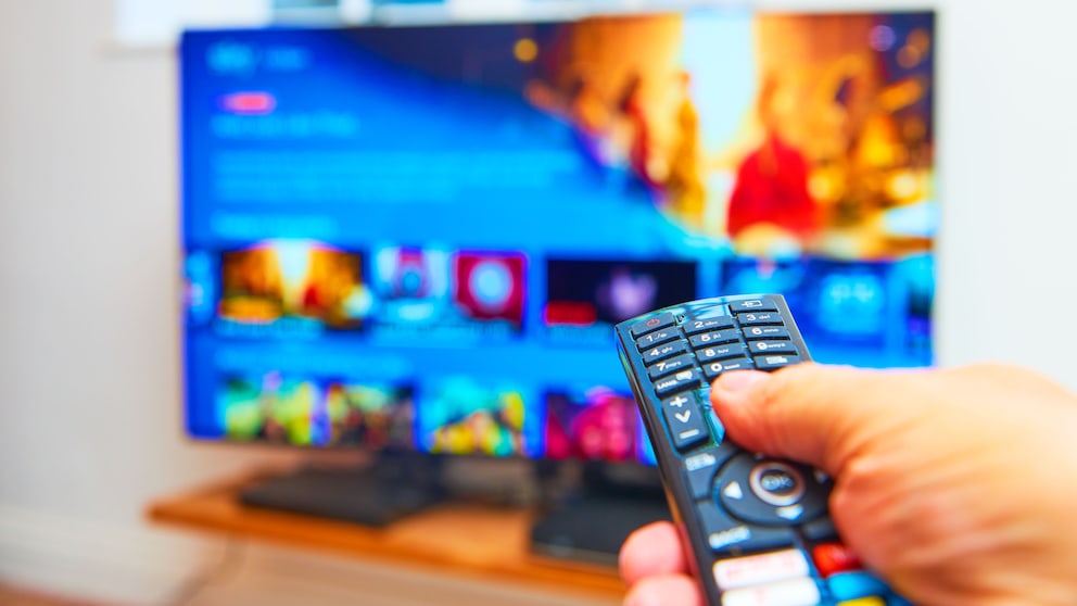 TV‑Fernbedienung im Vordergrund, unscharf gestellter Fernseher im Hintergrund