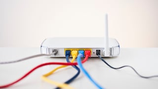 Weißer WLAN-Router auf einer hellen Oberfläche vor neutralem Hintergrund. Auf der Rückseite sind mehrere Anschlüsse mit eingesteckten Netzwerkkabeln in Blau, Gelb, Hellblau und Rot zu sehen. Rechts steht eine einzelne weiße Antenne, daneben ist ein schwarzes Kabel angeschlossen. Die Kabel verlaufen nach vorn aus dem Bild und sind teilweise unscharf dargestellt.