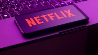 Alt-Text: Ein Smartphone mit rotem Netflix-Schriftzug liegt schräg auf der Tastatur eines Laptops. Das Bild ist in violett-pinkem Licht aufgenommen.