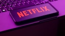 Alt-Text: Ein Smartphone mit rotem Netflix-Schriftzug liegt schr&auml;g auf der Tastatur eines Laptops. Das Bild ist in violett-pinkem Licht aufgenommen.