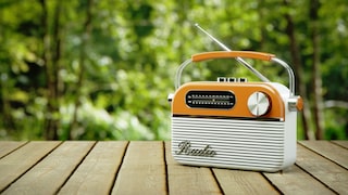 Orangefarbenes tragbares Radio mit Tragegriff und ausgezogener Antenne auf einem Holztisch. Auf der Vorderseite sind eine analoge AM- und FM-Frequenzskala, ein großer Drehknopf und mehrere Bedienelemente sichtbar. Im Hintergrund sind unscharfe grüne Bäume und Pflanzen zu sehen.