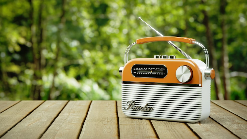 Orangefarbenes tragbares Radio mit Tragegriff und ausgezogener Antenne auf einem Holztisch. Auf der Vorderseite sind eine analoge AM- und FM-Frequenzskala, ein großer Drehknopf und mehrere Bedienelemente sichtbar. Im Hintergrund sind unscharfe grüne Bäume und Pflanzen zu sehen.