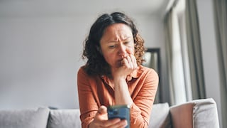 Person sitzt auf einem hellen Sofa in einem Wohnzimmer und hält ein Smartphone in der rechten Hand. Die Person beugt sich nach vorne und stützt das Kinn auf die linke Hand. Im Hintergrund sind ein Fenster mit Vorhängen und ein Spiegel zu sehen.