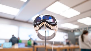 Apple Vision Pro auf einer Metallhalterung in einem Apple Store. Die Datenbrille ist mittig im Bild zu sehen, im Hintergrund stehen mehrere Personen zwischen Holztischen. Der Verkaufsraum ist hell beleuchtet, die Umgebung erscheint unscharf. Ein Kabel führt von der Brille zu einem Gerät auf dem Tisch.