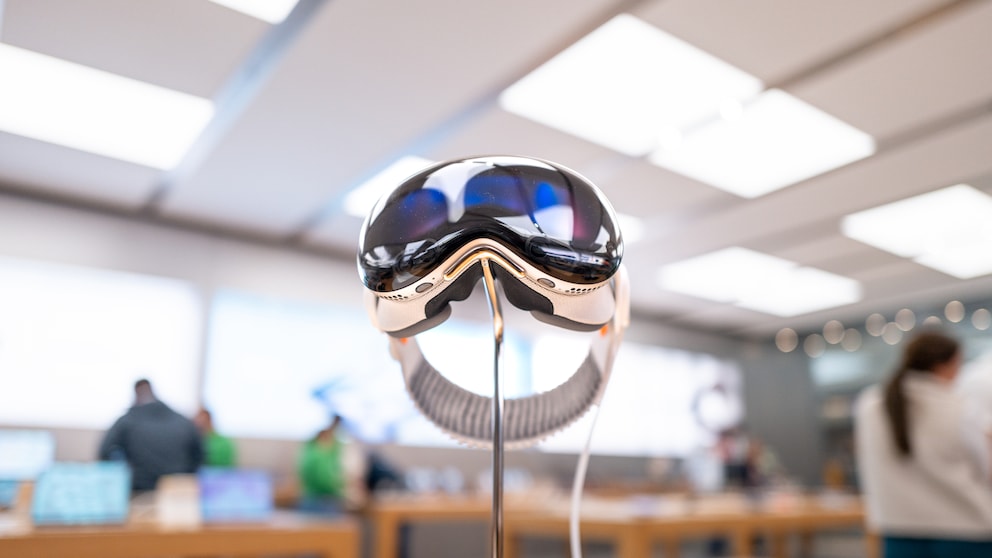 Apple Vision Pro auf einer Metallhalterung in einem Apple Store. Die Datenbrille ist mittig im Bild zu sehen, im Hintergrund stehen mehrere Personen zwischen Holztischen. Der Verkaufsraum ist hell beleuchtet, die Umgebung erscheint unscharf. Ein Kabel führt von der Brille zu einem Gerät auf dem Tisch.