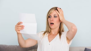 Person sitzt auf einem Sofa und hält ein Blatt Papier in der Hand. Eine Hand liegt am Kopf. Im Hintergrund ist eine helle Wand zu sehen. Das Dokument wird aufmerksam betrachtet.