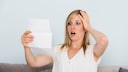 Person sitzt auf einem Sofa und h&auml;lt ein Blatt Papier in der Hand. Eine Hand liegt am Kopf. Im Hintergrund ist eine helle Wand zu sehen. Das Dokument wird aufmerksam betrachtet.