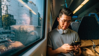 Person sitzt in einem Zug auf einem Fensterplatz und hält ein Smartphone in den Händen. Um den Hals liegen weiße Kopfhörer. Im Fenster spiegelt sich die Person, während draußen Gebäude und Bahnanlagen vorbeiziehen. Auf dem Nebensitz liegt ein heller Rucksack.