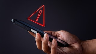 Eine Hand hält ein schwarzes Smartphone vor dunklem Hintergrund. Über dem Gerät schwebt ein rotes Warnsymbol in Form eines leuchtenden Dreiecks mit Ausrufezeichen. Das Motiv steht für eine Sicherheitswarnung oder ein mögliches Risiko im Zusammenhang mit Smartphones.