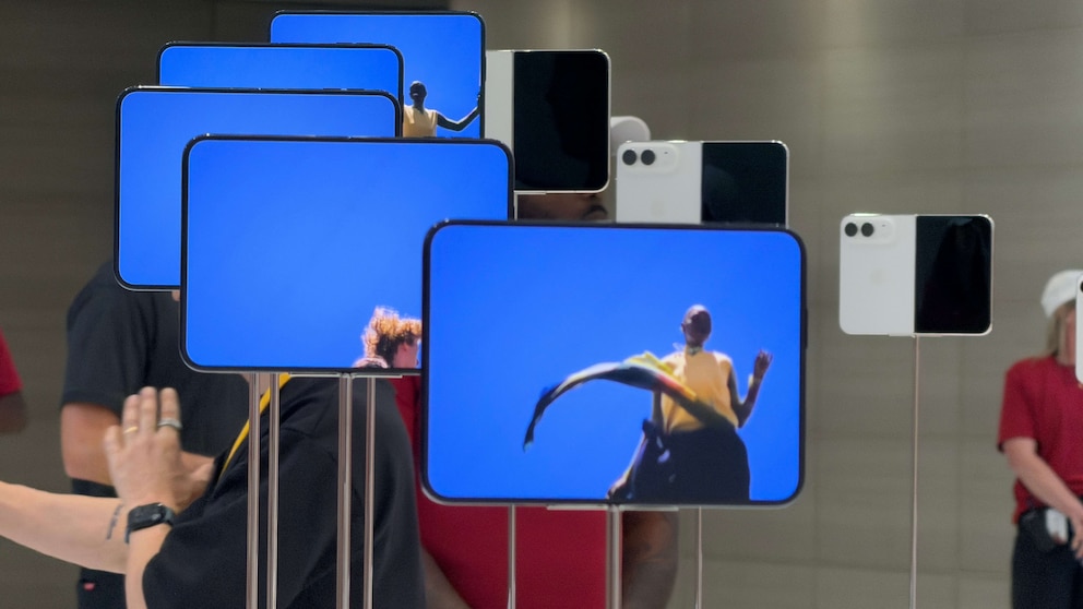 Mehrere aufgeklappte iPhone-Duo-Smartphones stehen auf schmalen Halterungen. Die Ger&auml;te zeigen ein gro&szlig;es blaues Display mit einer Person im Bild. Daneben sind einige Modelle zusammengeklappt zu sehen. Im Hintergrund befinden sich Besucher und ein heller Ausstellungsbereich.