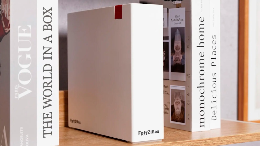 Wei&szlig;e FritzBox DSL/Fiber 7-90 steht auf einem Holzregal zwischen mehreren B&uuml;chern. Das Ger&auml;t besitzt ein schlichtes rechteckiges Design mit glatter Front und einem roten Element an der oberen Kante. Auf der Vorderseite ist das Fritz!Box-Logo zu sehen. Links und rechts neben dem Router stehen B&uuml;cher mit den Titeln &bdquo;VOGUE&ldquo;, &bdquo;THE WORLD IN A BOX&ldquo;, &bdquo;monochrome home&ldquo; und &bdquo;Delicious Places&ldquo;.