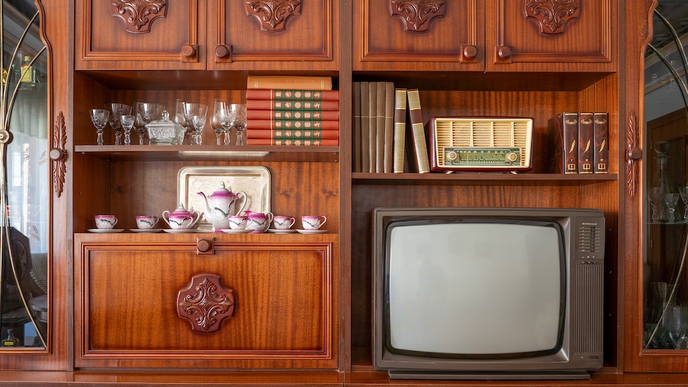 Alter Röhrenfernseher steht in einer dunklen Holzschrankwand. Daneben befinden sich Bücher, Gläser, ein Teeservice und ein Radio.