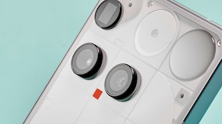 Rückseite eines weißen Nothing-Smartphones in schräger Ansicht. Sichtbar sind drei große runde Kameramodule, ein kleiner Sensor sowie die für Nothing typische transparente Gestaltung mit geometrischen Elementen unter der Rückseite. Das Gerät liegt vor einem helltürkisen Hintergrund.