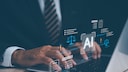 Ein Mann in Anzug sitzt an einem Schreibtisch und arbeitet mithilfe von AI auf seinem Laptop.