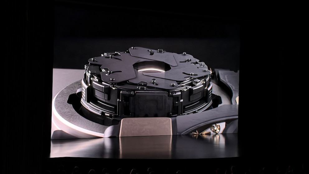 Gro&szlig;aufnahme einer kreisf&ouml;rmigen Kamerakomponente auf einer Pr&auml;sentationsfolie. Das Bauteil besteht aus mehreren &uuml;bereinanderliegenden schwarzen Metallteilen mit Schrauben und beweglichen Segmenten rund um eine zentrale &Ouml;ffnung. Die technische Darstellung ist vor dunklem Hintergrund zu sehen.