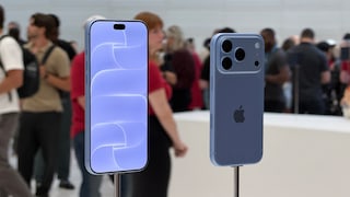 Zwei blaue Apple-Smartphones stehen auf Metallhalterungen in einem hellen Ausstellungsraum. Links ist die Vorderseite mit eingeschaltetem Display zu sehen, rechts die Rückseite mit drei Kameralinsen. Im unscharfen Hintergrund befinden sich mehrere Personen und weitere Ausstellungsflächen.