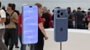 Zwei blaue Apple-Smartphones stehen auf Metallhalterungen in einem hellen Ausstellungsraum. Links ist die Vorderseite mit eingeschaltetem Display zu sehen, rechts die R&uuml;ckseite mit drei Kameralinsen. Im unscharfen Hintergrund befinden sich mehrere Personen und weitere Ausstellungsfl&auml;chen.