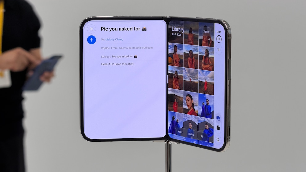 An unfolded iPhone Duo stands on a metal holder. On the left half of the screen, an email app is open, and on the right side, a photo library with several preview images is visible. Additional control elements are located on the right edge of the display. In the background, a person with a smartphone is seen out of focus.