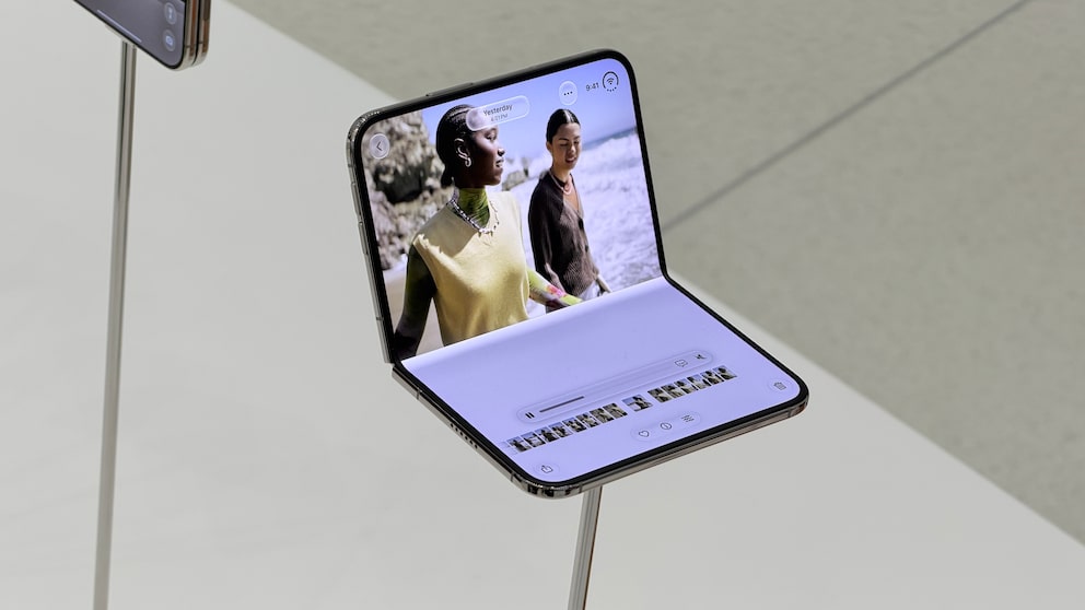 An unfolded iPhone Duo stands on a narrow metal holder. On the large inner display, an app with a video or photo of two people is visible. At the bottom of the screen, there are control elements and a bar with preview images. The device is on a white exhibition table in front of a bright background.