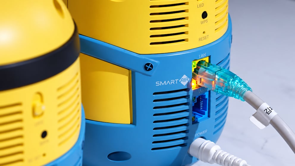 Close-up of the back of a yellow-blue router in Minion design. A network cable is plugged into a port labeled 'LAN.' Below are additional ports and a plugged-in power cable. The labels 'LED,' 'WPS,' 'RESET,' and 'POWER' are visible on the casing. Another router of the same design is partially visible and out of focus on the left foreground. The indicator lights on the network port are glowing green and orange.