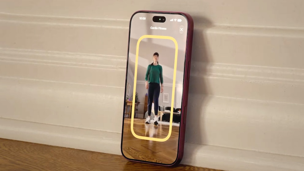 Ein Smartphone mit rotem Geh&auml;use lehnt an einer wei&szlig;en Wand auf einem Holzboden. Auf dem Display ist die Funktion &bdquo;Cardio Fitness&ldquo; zu sehen. Die Kamera erfasst eine Person, die in einem Raum steht. Um die Person herum wird ein gelber Rahmen angezeigt.