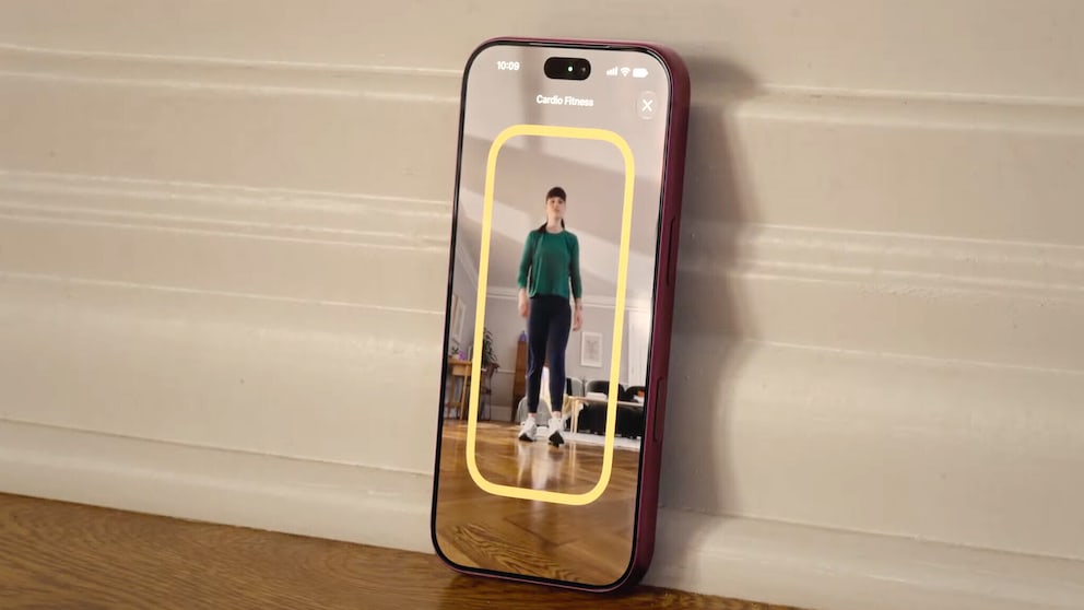 A smartphone with a red case leaning against a white wall on a wooden floor. The display shows the 'Cardio Fitness' function. The camera captures a person standing in a room. A yellow frame is displayed around the person.