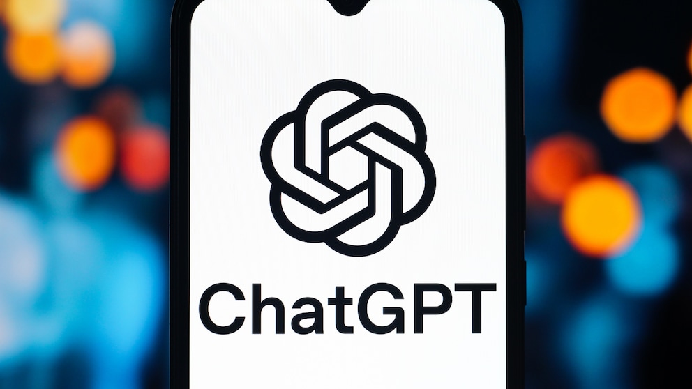Ein Smartphone steht hochkant vor einem unscharfen Hintergrund mit blauen, orangefarbenen und roten Lichtpunkten. Auf dem Display ist das schwarze ChatGPT-Logo auf weißem Hintergrund zu sehen. Unter dem Logo steht der Schriftzug „ChatGPT“. Am oberen Displayrand befindet sich eine kleine Aussparung für die Frontkamera. Das Smartphone nimmt den Großteil des Bildes ein und ist mittig platziert.