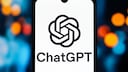 Ein Smartphone steht hochkant vor einem unscharfen Hintergrund mit blauen, orangefarbenen und roten Lichtpunkten. Auf dem Display ist das schwarze ChatGPT-Logo auf wei&szlig;em Hintergrund zu sehen. Unter dem Logo steht der Schriftzug &bdquo;ChatGPT&ldquo;. Am oberen Displayrand befindet sich eine kleine Aussparung f&uuml;r die Frontkamera. Das Smartphone nimmt den Gro&szlig;teil des Bildes ein und ist mittig platziert.